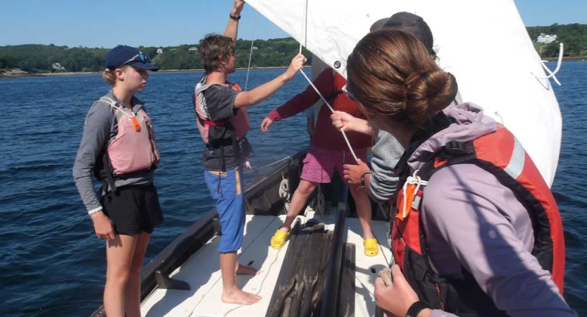 People wearing life jackets stand and work with the white sail of a sail boat. 